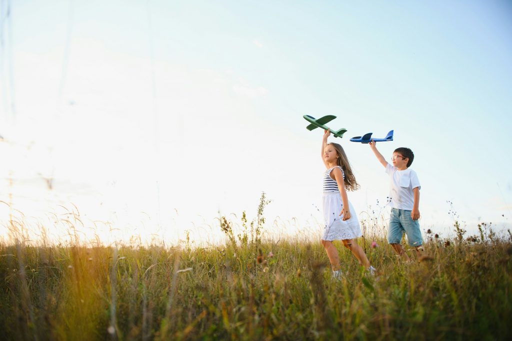 Running boy and girl holding two green and blue airplanes toy in the field during summer sunny day